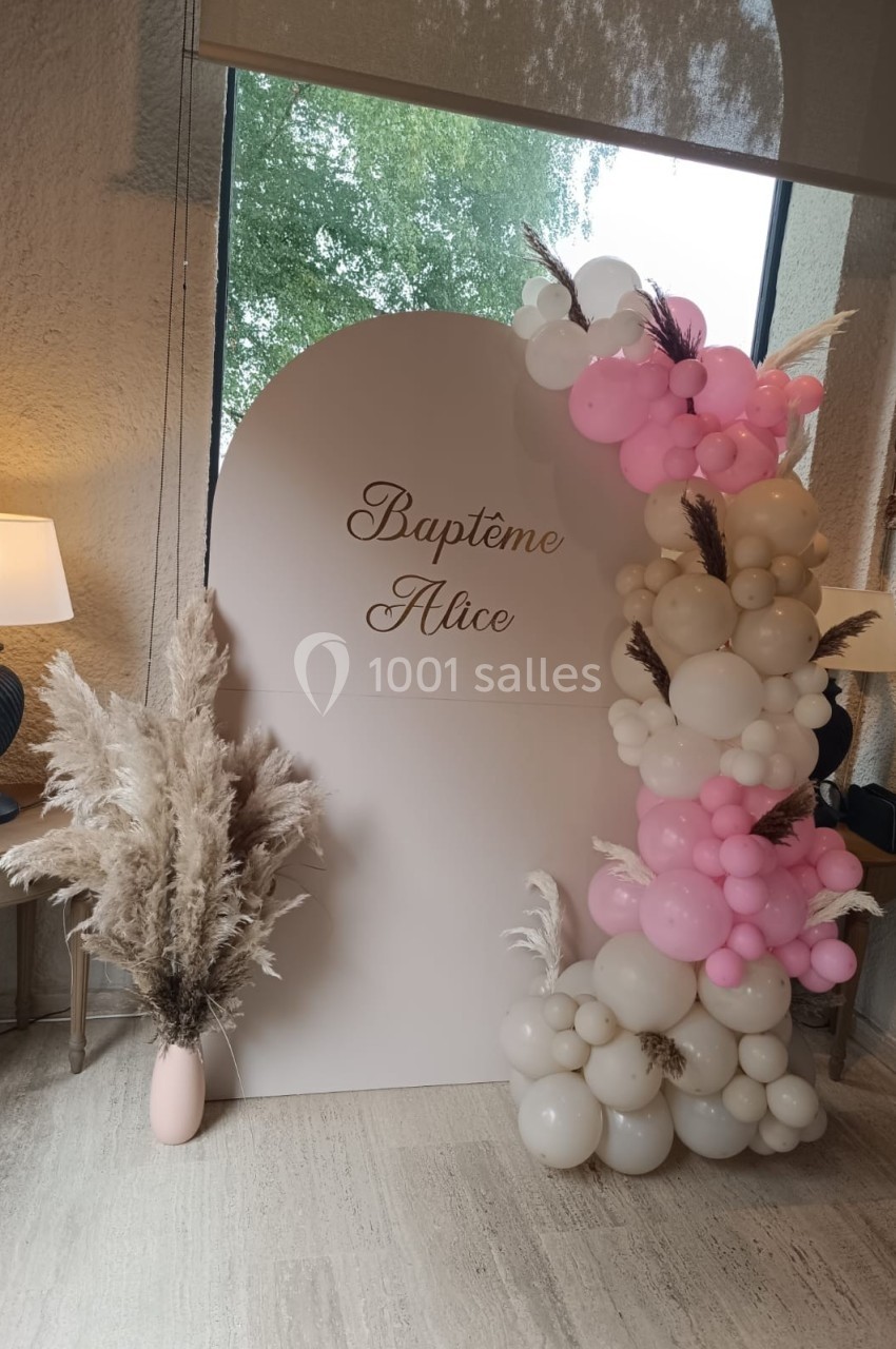 Mur décoratif avec inscription ’Baptême Alice’, ballons roses et blancs, pampas dans un vase, près d'une fenêtre.