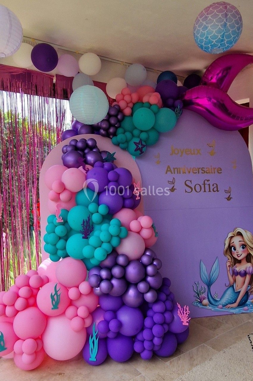 Décoration d'anniversaire sur le thème des sirènes avec des ballons colorés, une arche et une illustration de sirène.