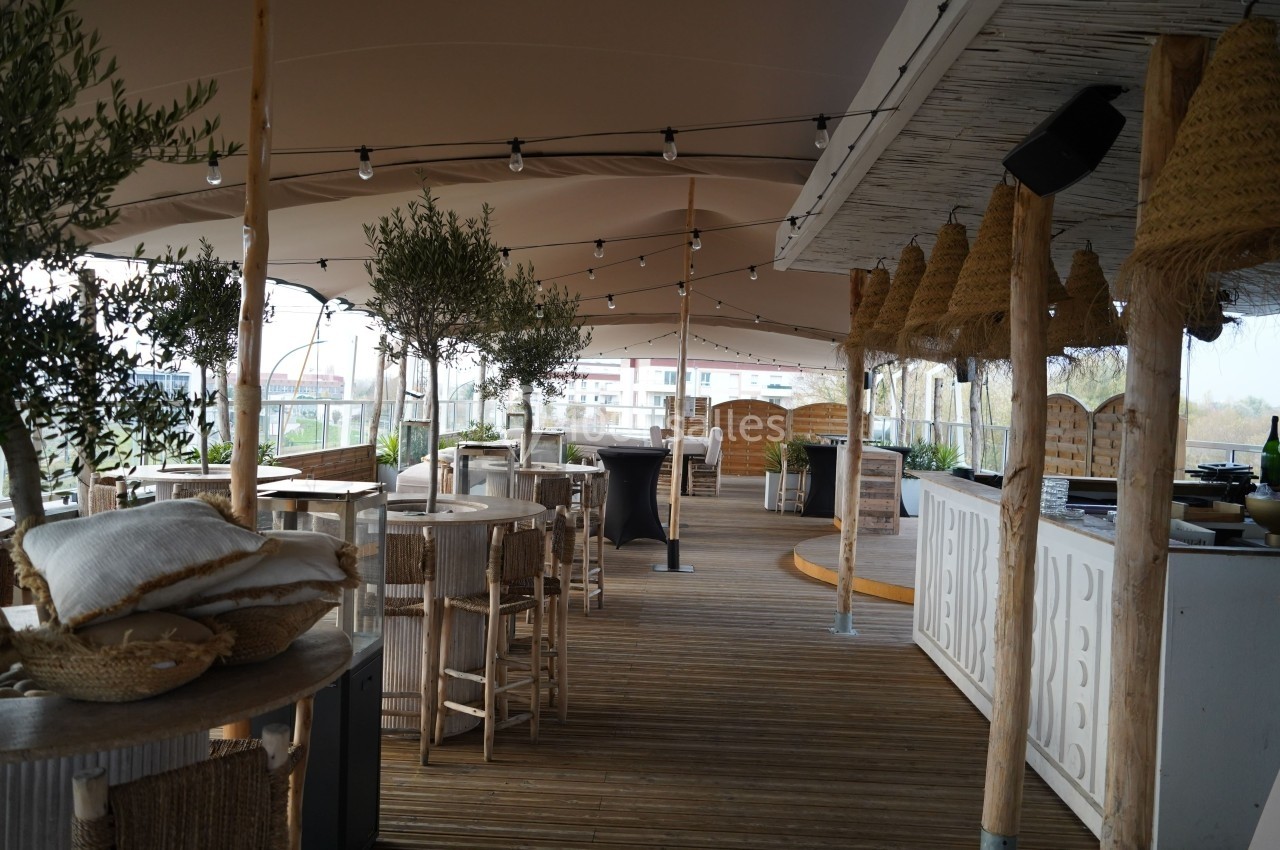Terrasse en bois aménagée avec tables, chaises, plantes et éclairage suspendu sous une grande toile tendue.