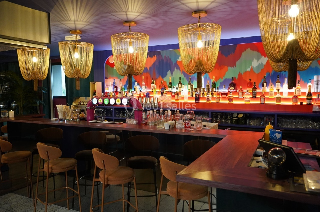 Bar coloré avec comptoir en bois, tabourets rembourrés, luminaires suspendus et étagères garnies de bouteilles.