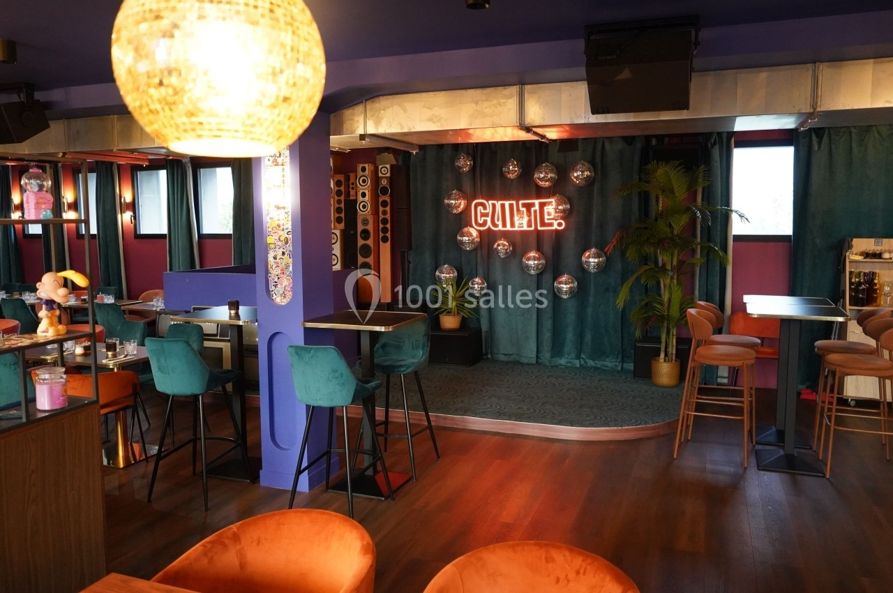 Salle de bar colorée avec une petite scène, des tabourets, des tables hautes et une enseigne lumineuse ’CULTE’.