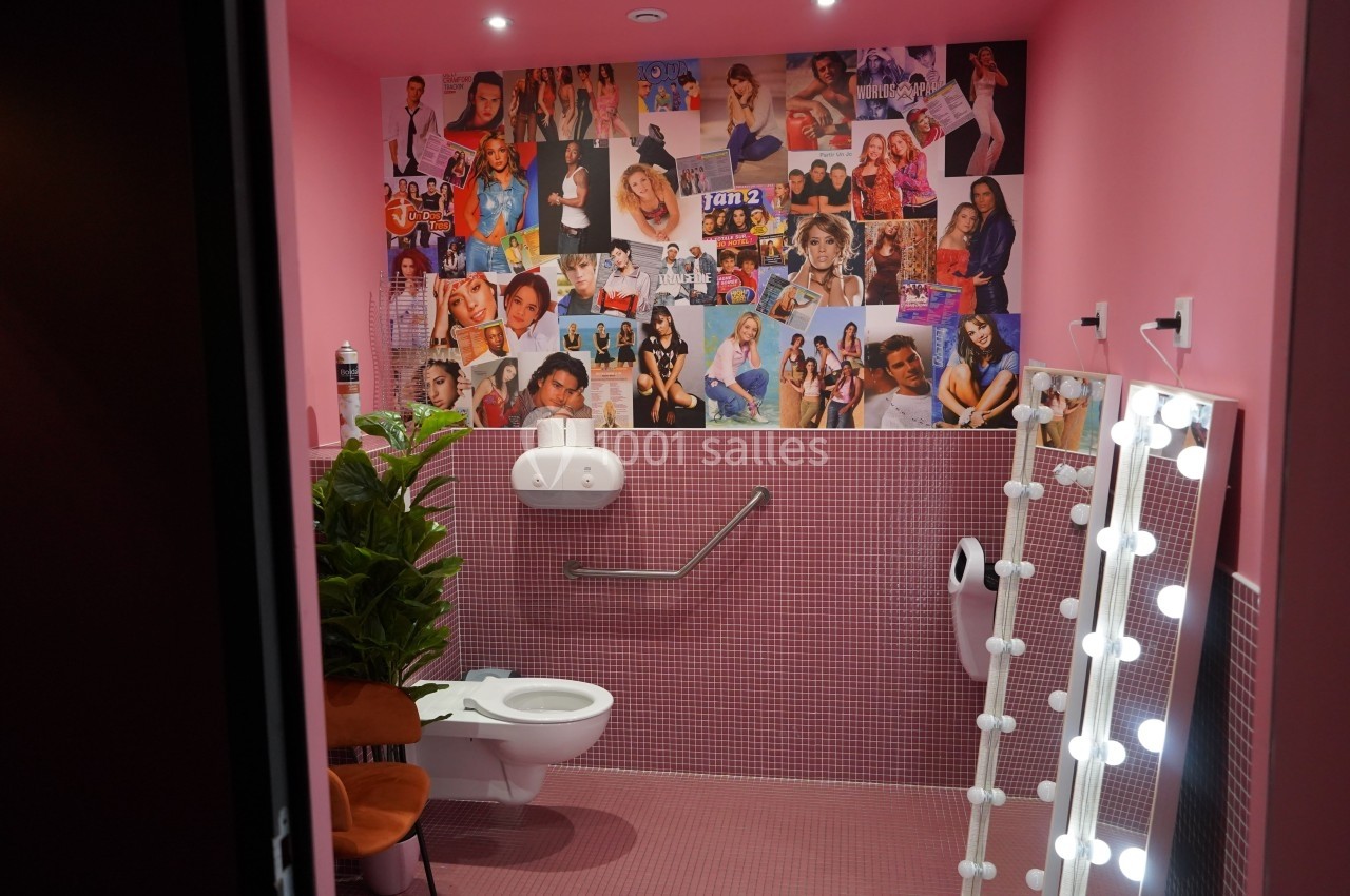 Toilettes roses décorées de mosaïques, affiches rétro sur les murs et miroir éclairé par des ampoules.