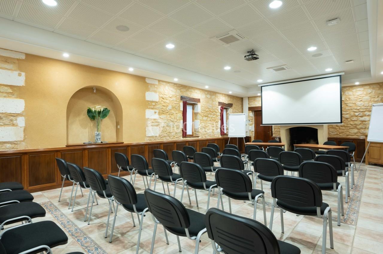 Salle de réunion avec des rangées de chaises noires, un écran de projection, des tableaux blancs et des murs en pierre.