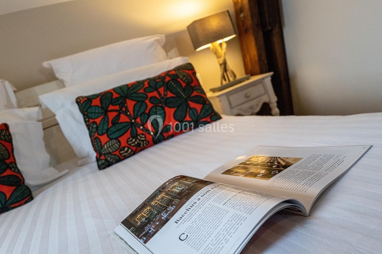 Lit avec draps blancs, coussins à motifs colorés, lampe allumée sur une table de chevet et magazine ouvert au premier plan.