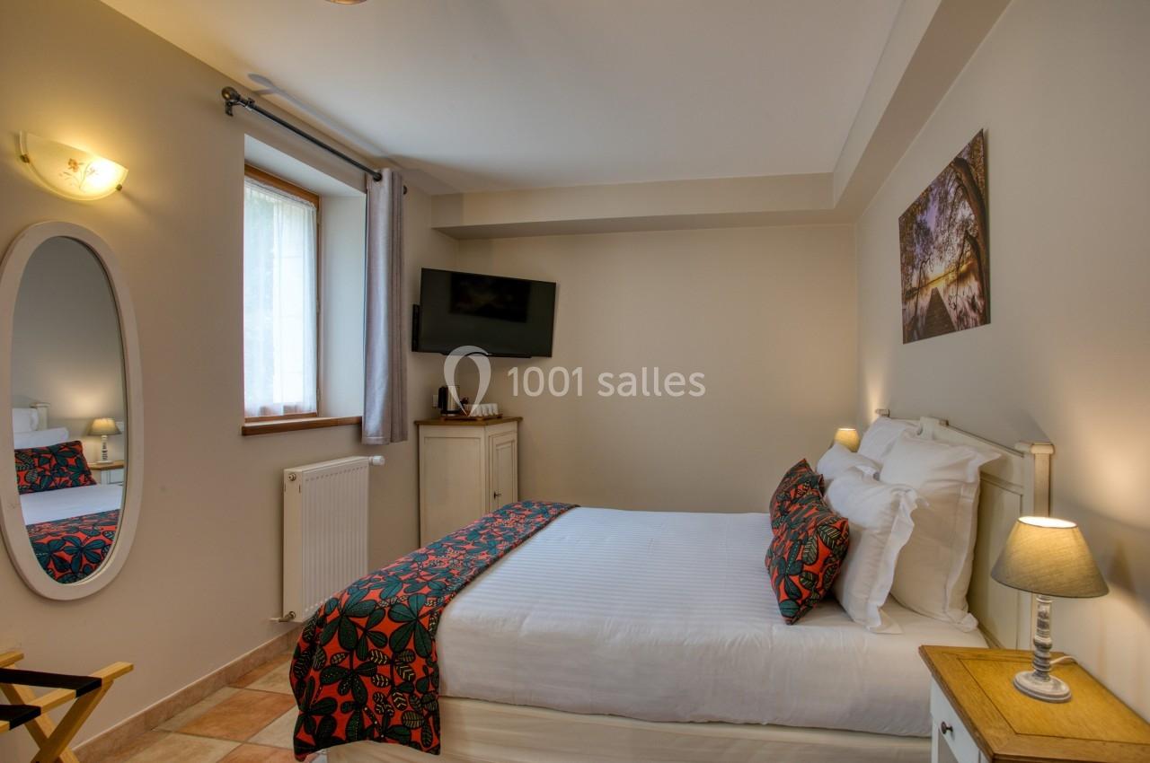 Chambre lumineuse avec lit double, décoration sobre, télévision murale, miroir ovale et fenêtre avec rideaux.