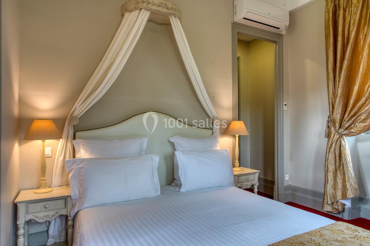 Lit double avec baldaquin, draps blancs, tables de chevet avec lampes allumées, rideaux dorés et mur beige.