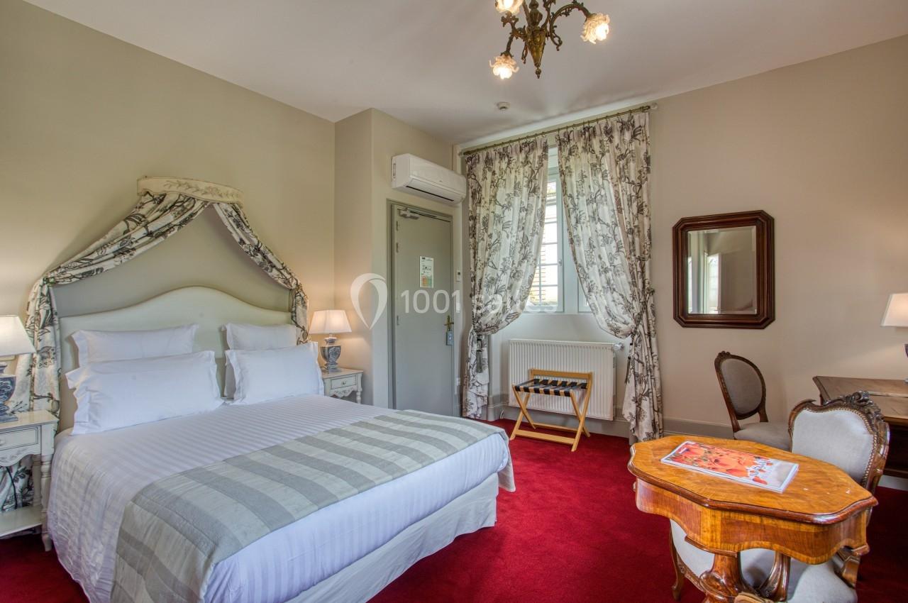 Chambre élégante avec lit double, mobilier classique, rideaux imprimés, moquette rouge et éclairage chaleureux.