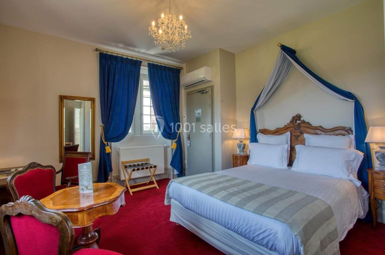 Chambre élégante avec lit à baldaquin, rideaux bleus, mobilier en bois et moquette rouge, éclairée par un lustre.
