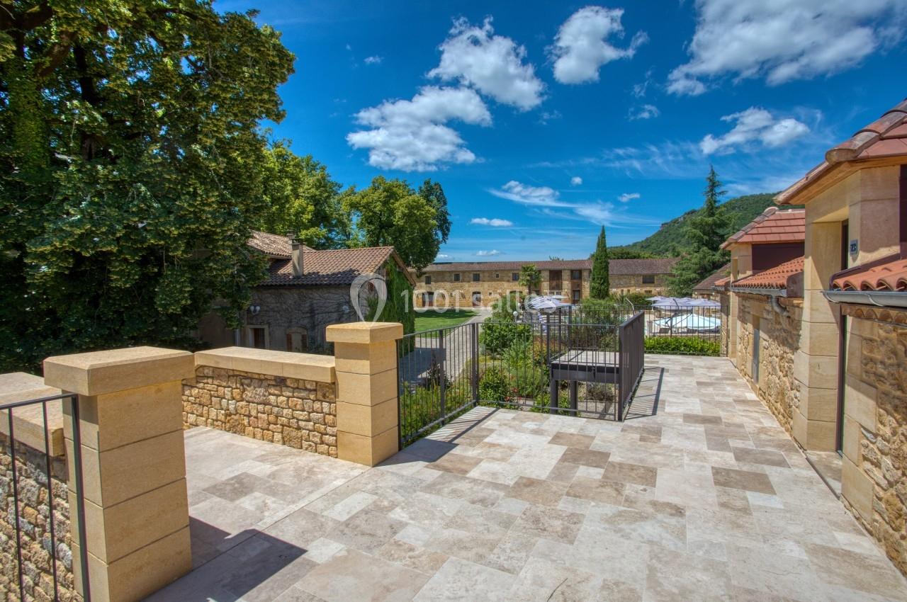 Terrasse en pierre avec vue sur un jardin verdoyant, des bâtiments en pierre et un ciel bleu parsemé de nuages.