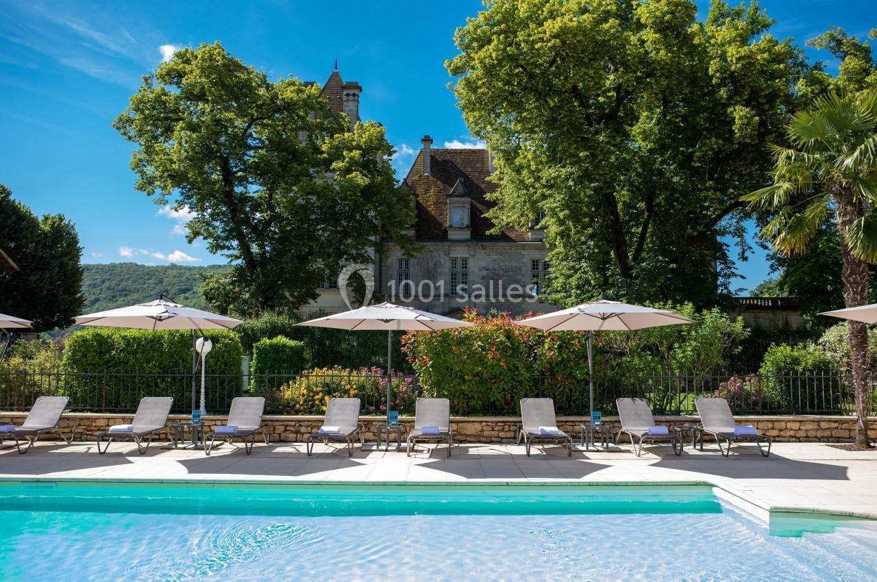 Piscine extérieure entourée de chaises longues et parasols, avec un manoir et des arbres en arrière-plan.