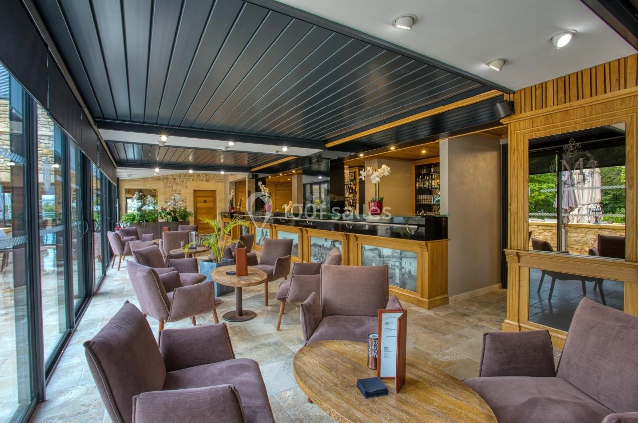 Espace lounge moderne avec fauteuils, tables basses, bar en bois et grandes baies vitrées donnant sur l'extérieur.