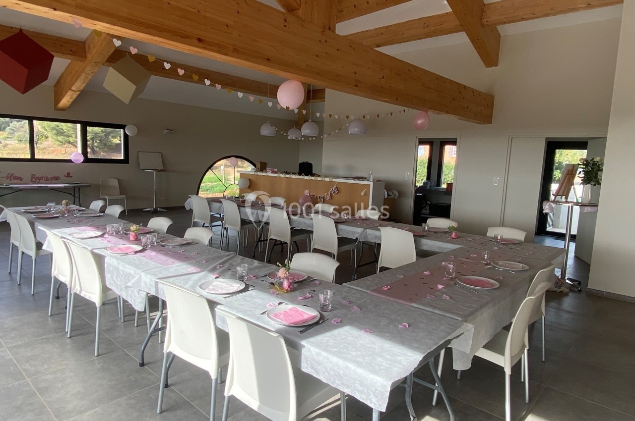 Salle décorée pour un événement, avec tables alignées, nappes grises, assiettes roses et guirlandes suspendues.
