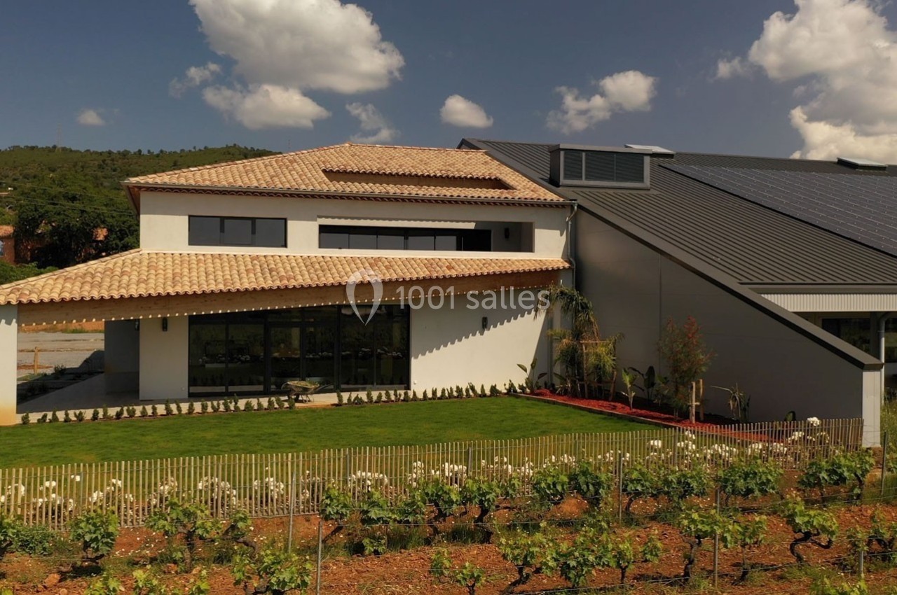 Bâtiment moderne avec toit en tuiles et panneaux solaires, entouré de vignes et d'un jardin verdoyant sous un ciel bleu.