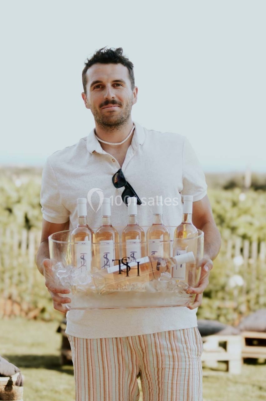 Un homme tenant un seau transparent rempli de bouteilles de vin rosé, en extérieur avec un fond flou de verdure.