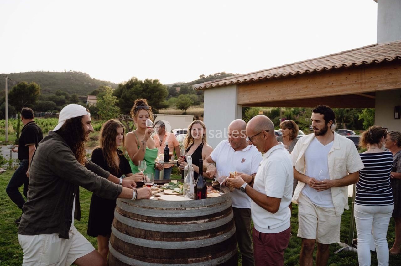 Un groupe de personnes partage un apéritif autour d'un tonneau en extérieur, près d'une maison.