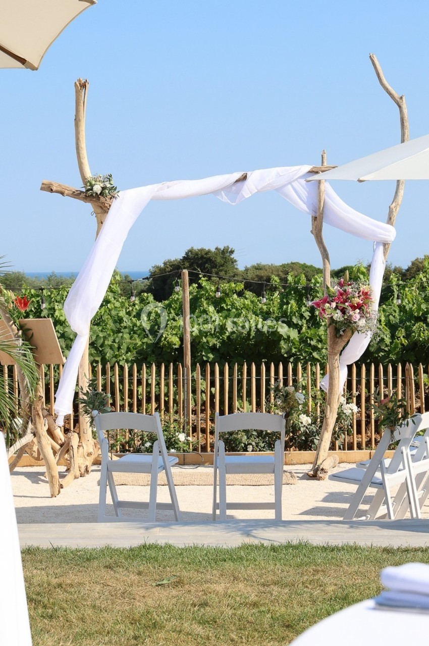 Arche de mariage décorée de tissu blanc et de fleurs, installée en extérieur face à des vignes et un ciel dégagé.