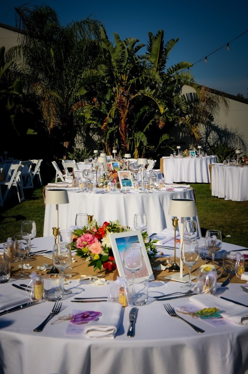 Tables rondes décorées pour un événement en extérieur, avec nappes blanches, fleurs et lampes, sous un ciel dégagé.