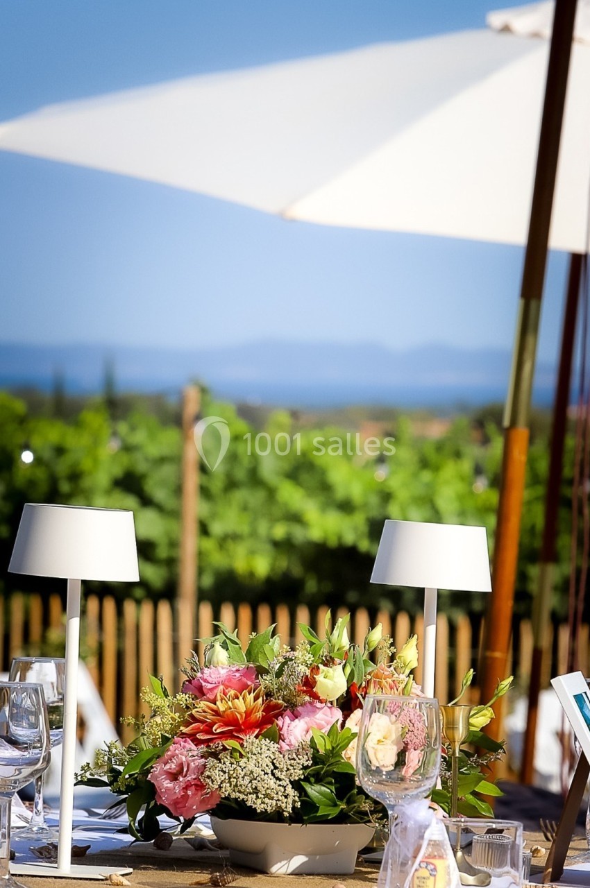 Centre de table floral avec des dahlias et des verres, en extérieur sous un parasol avec vue sur un paysage ensoleillé.