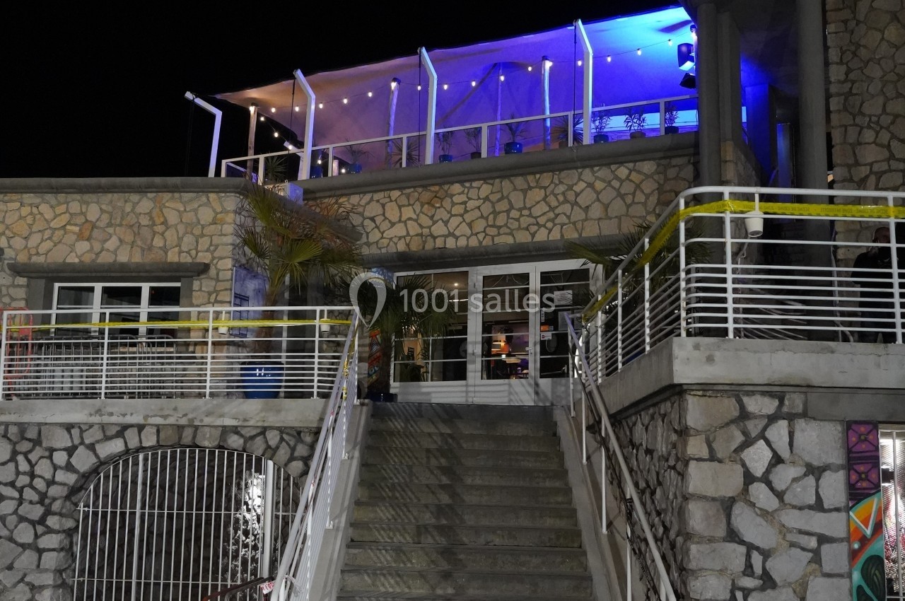 Façade d'un bâtiment en pierre avec terrasse éclairée en bleu, accessible par un escalier central.