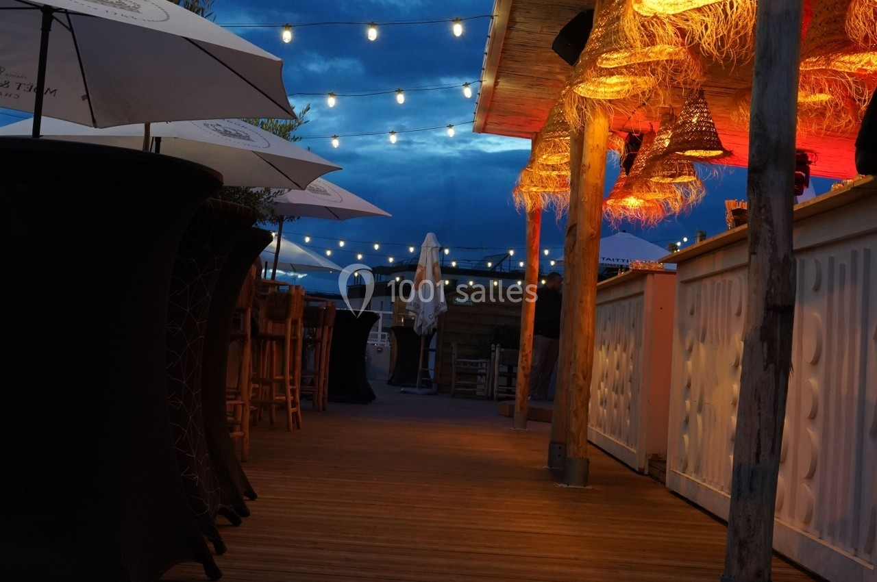 Terrasse en soirée avec guirlandes lumineuses, parasols, mobilier en bois et éclairage chaleureux sous un ciel sombre.