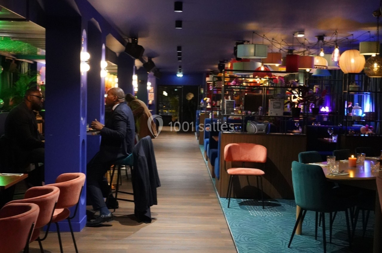 Salle de restaurant moderne avec éclairage tamisé, chaises colorées et clients discutant à des tables et au bar.