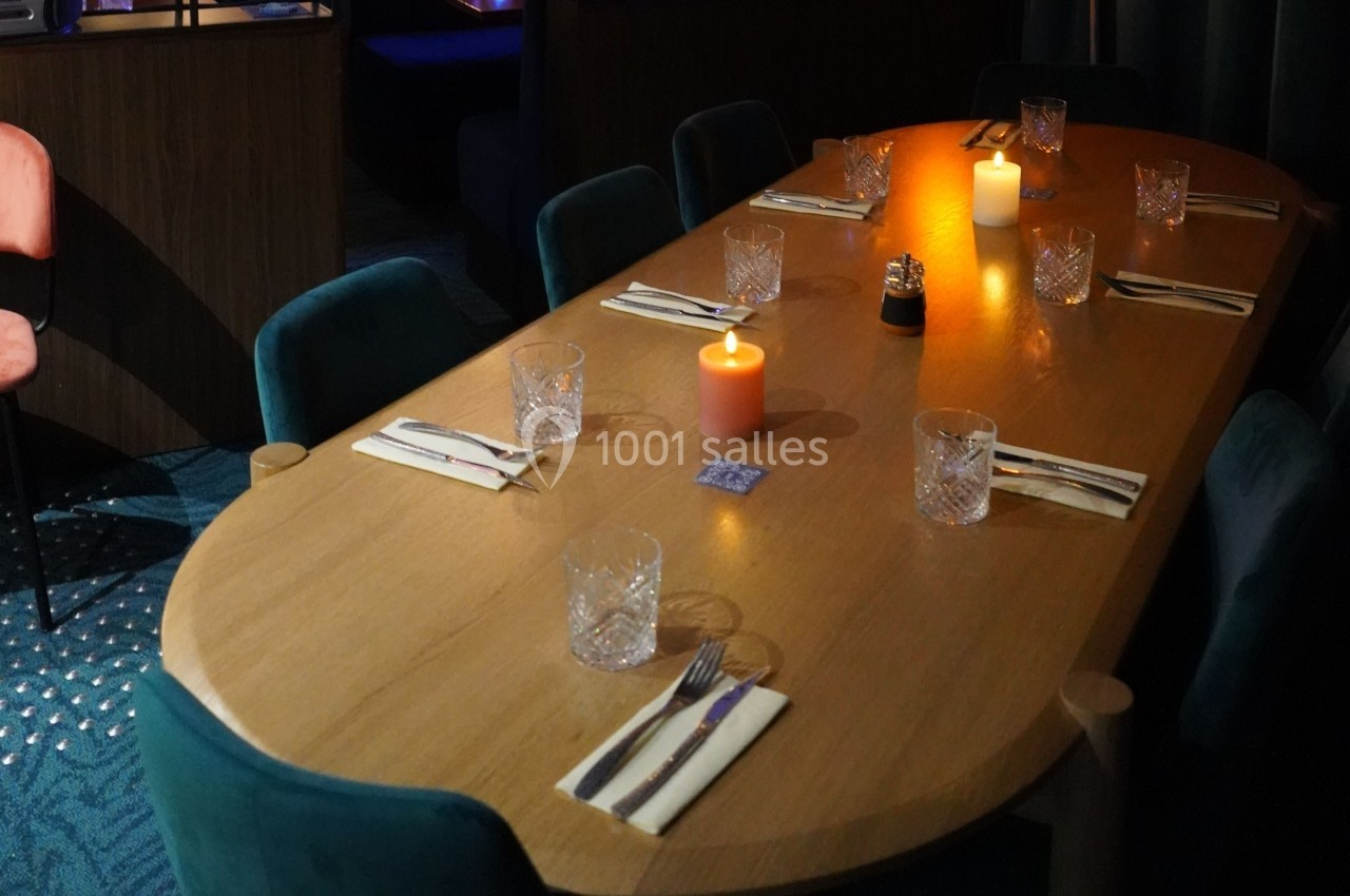 Table en bois ovale dressée pour un repas, avec bougies allumées, verres, couverts et chaises rembourrées.