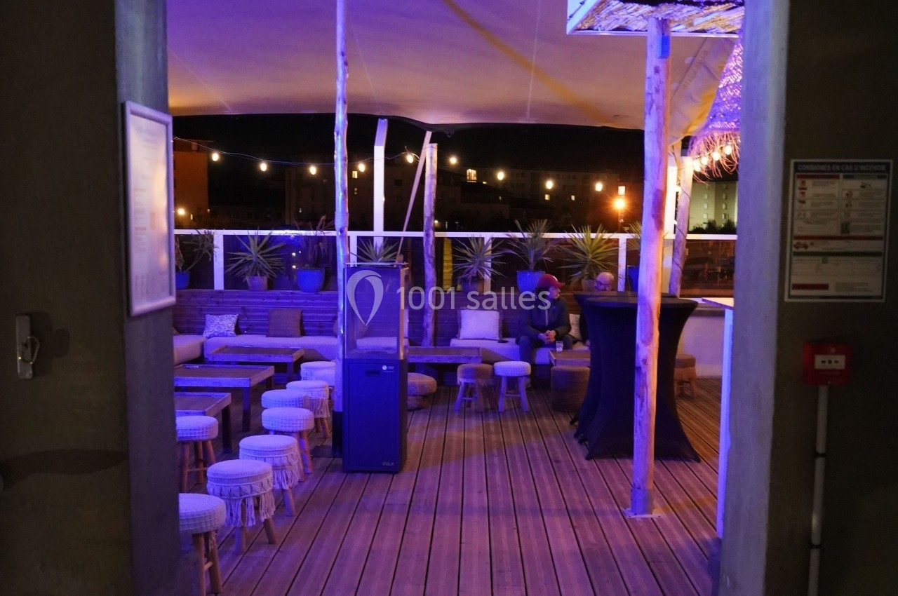 Terrasse extérieure éclairée de nuit avec tables, tabourets et plantes, ambiance chaleureuse et décor moderne.