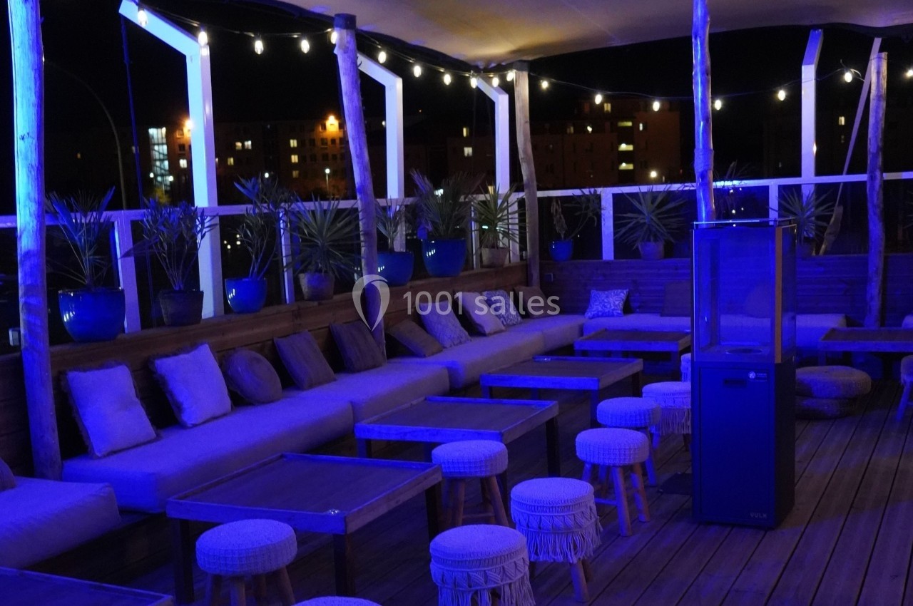 Terrasse extérieure éclairée en soirée, avec tables basses, tabourets, banquettes et plantes en pots sous des guirlandes…