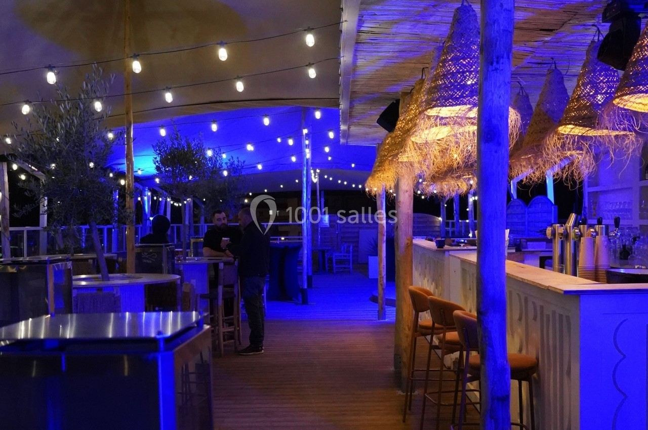 Terrasse extérieure éclairée de guirlandes lumineuses, avec un bar en bois, des tabourets et quelques personnes présentes.