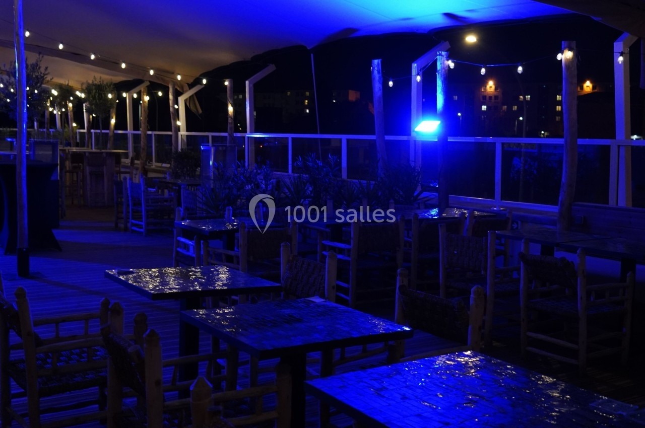 Terrasse extérieure éclairée par des lumières bleues et guirlandes lumineuses, avec des tables et chaises en bois.