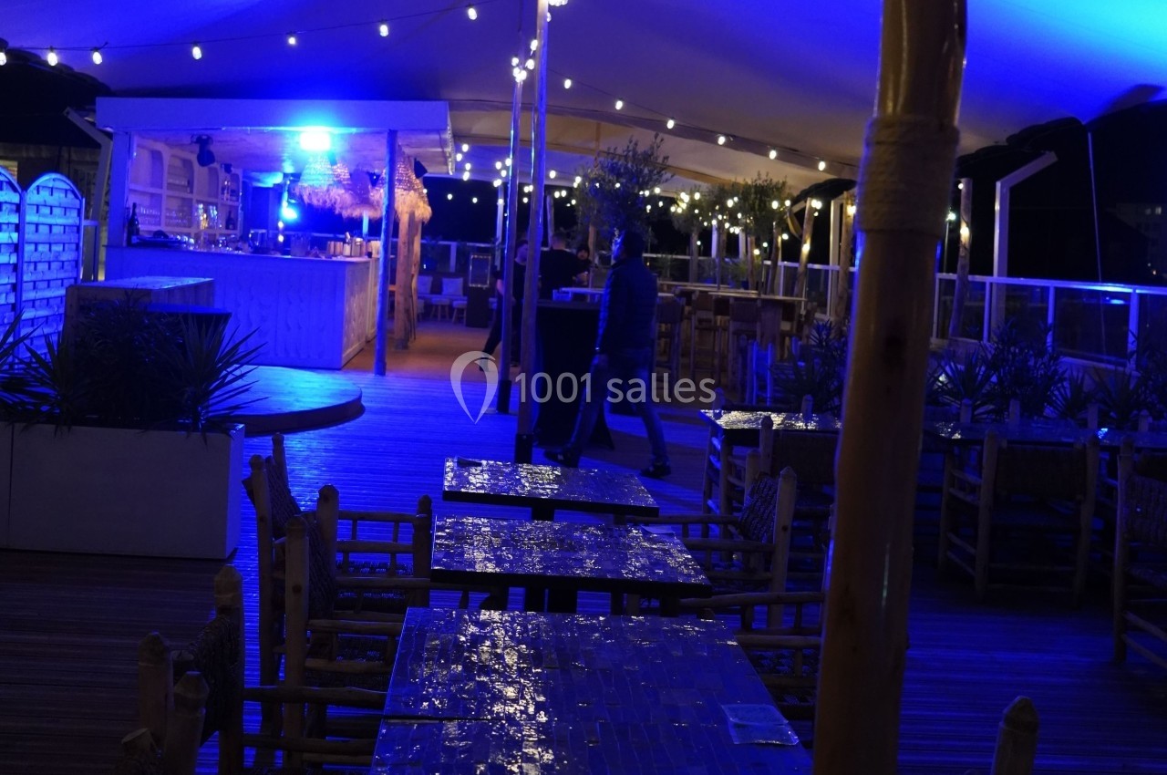 Terrasse extérieure éclairée de nuit avec tables en bois, guirlandes lumineuses et un bar en arrière-plan.