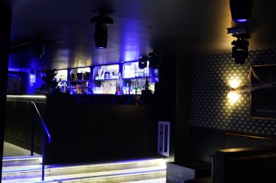 Bar moderne avec éclairage bleu, étagères remplies de bouteilles et ambiance tamisée.