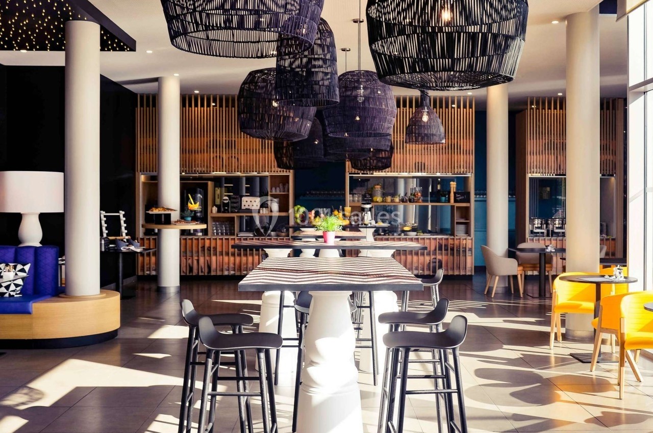 Salle de restaurant moderne avec tables hautes, chaises noires et suspensions en osier noir, éclairée par la lumière…