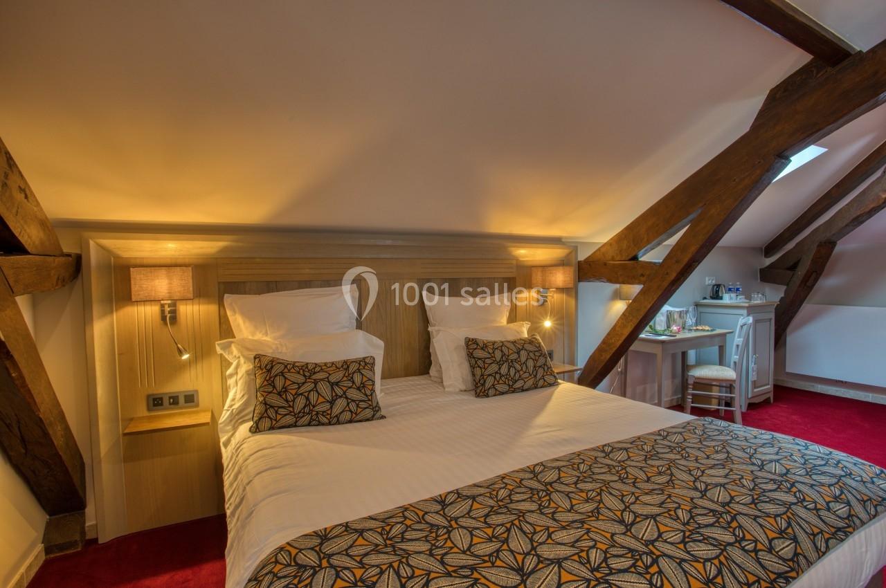 Chambre mansardée avec lit double, poutres apparentes, éclairage chaleureux et coin bureau sur moquette rouge.