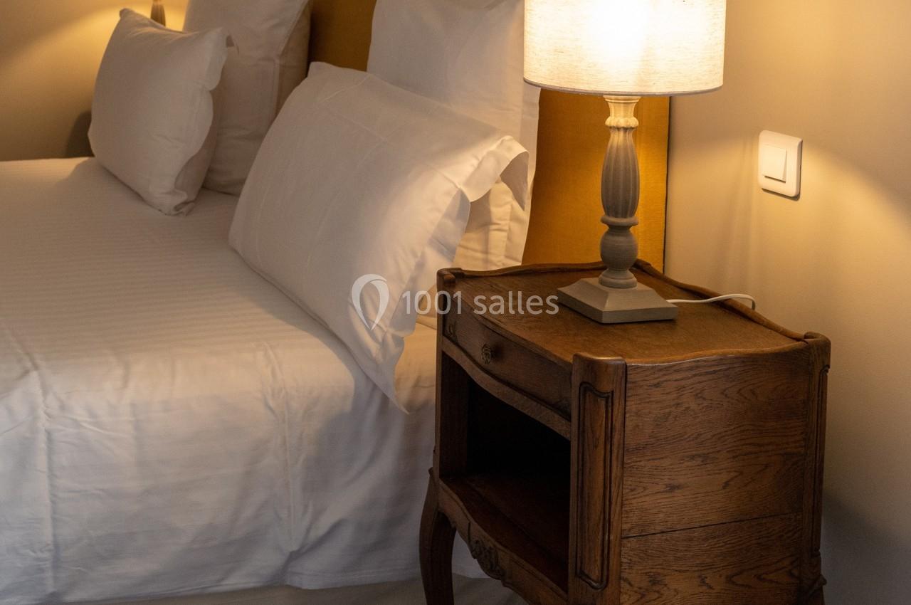 Table de chevet en bois avec lampe allumée, placée à côté d'un lit avec tête de lit jaune et draps blancs.