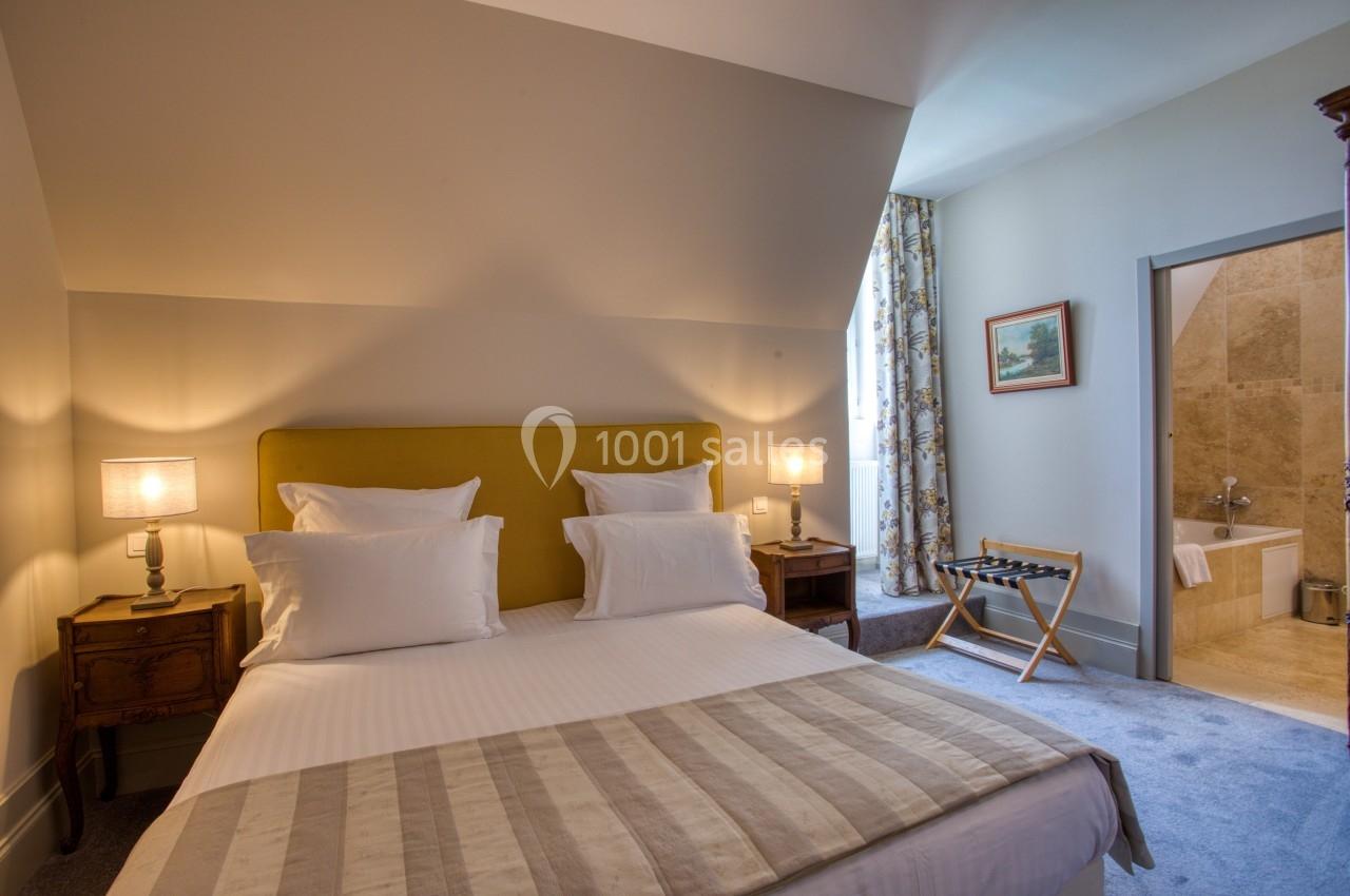 Chambre lumineuse avec lit double, tables de chevet, lampes, fauteuil pliant et salle de bain attenante.