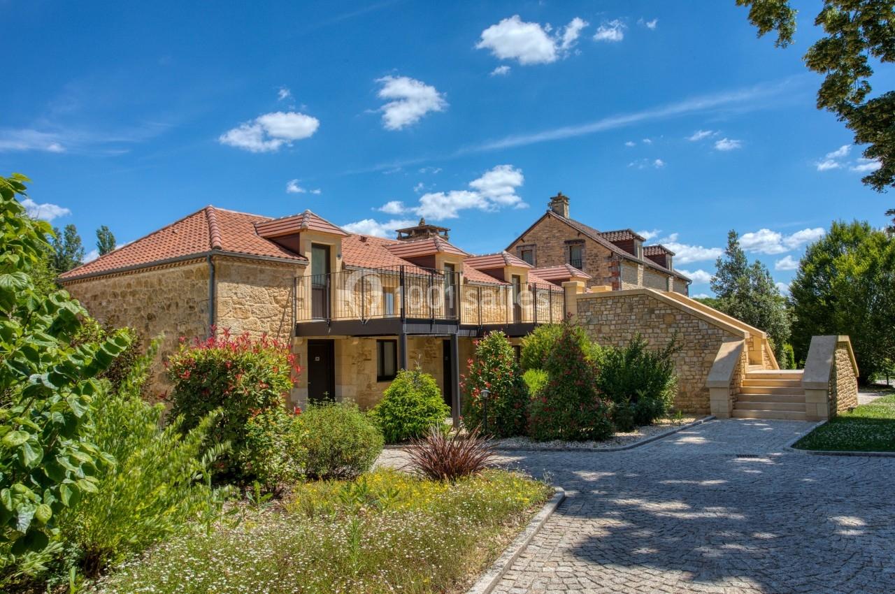 Maison en pierre avec toit en tuiles rouges, entourée de végétation et d'une allée pavée sous un ciel bleu.