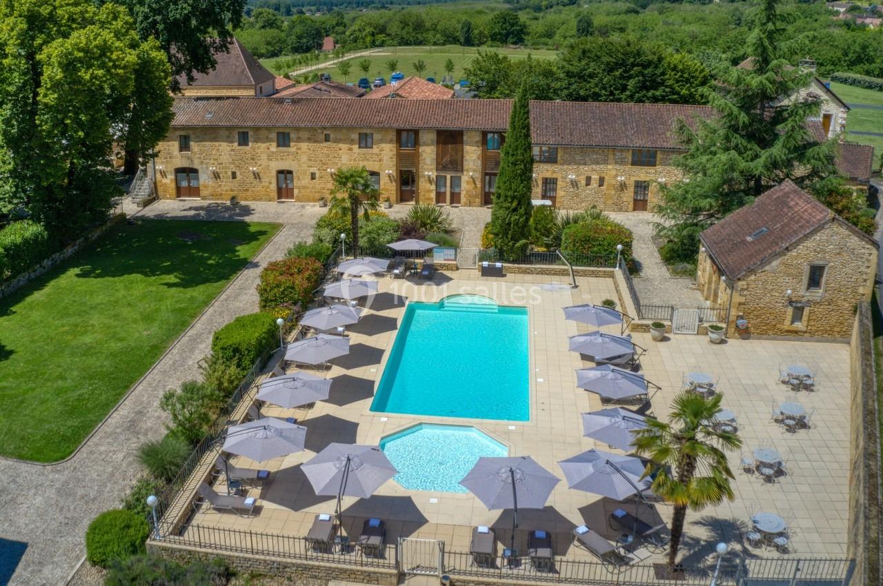 Vue aérienne d'une piscine entourée de chaises longues et parasols, située dans une cour avec bâtiments en pierre et espaces…