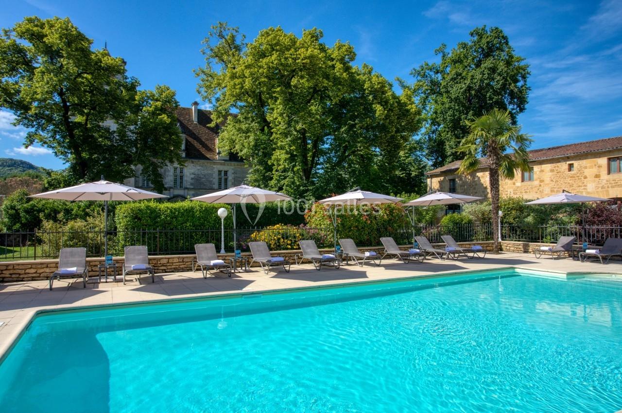 Piscine extérieure entourée de transats et parasols, avec des arbres et des bâtiments en pierre en arrière-plan.