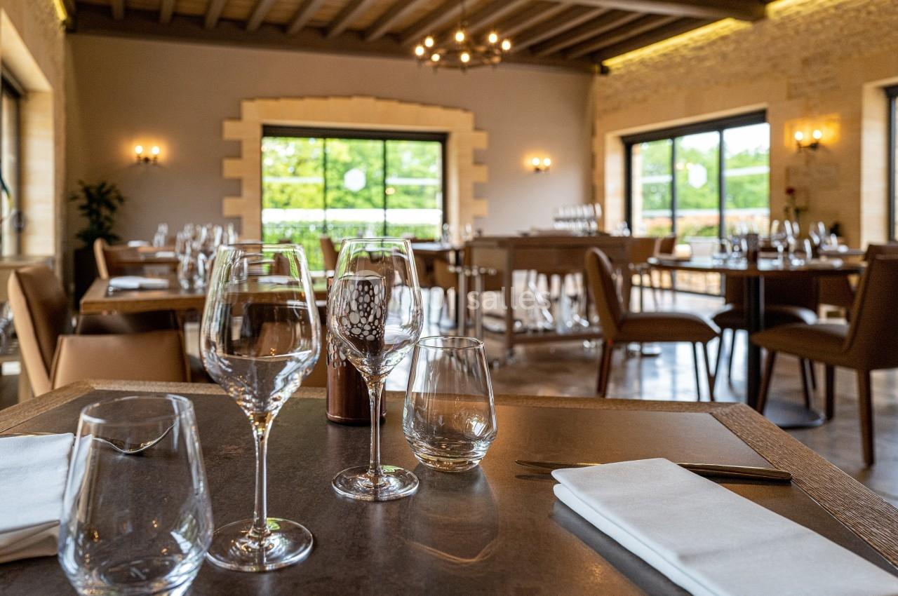 Salle de restaurant élégante avec tables dressées, verres et serviettes, éclairée par des fenêtres donnant sur un jardin.