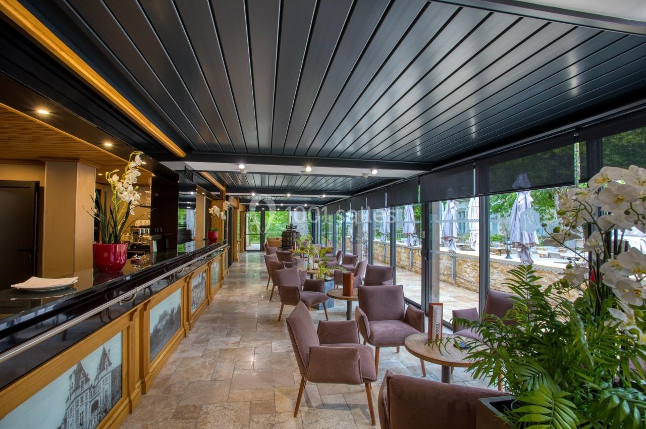 Intérieur d'une terrasse couverte avec mobilier en bois et tissu, grandes baies vitrées et vue sur un espace extérieur.