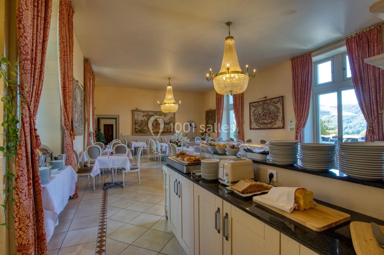 Salle lumineuse avec tables dressées, lustres, rideaux rouges et buffet comprenant vaisselle, pain et grille-pain.