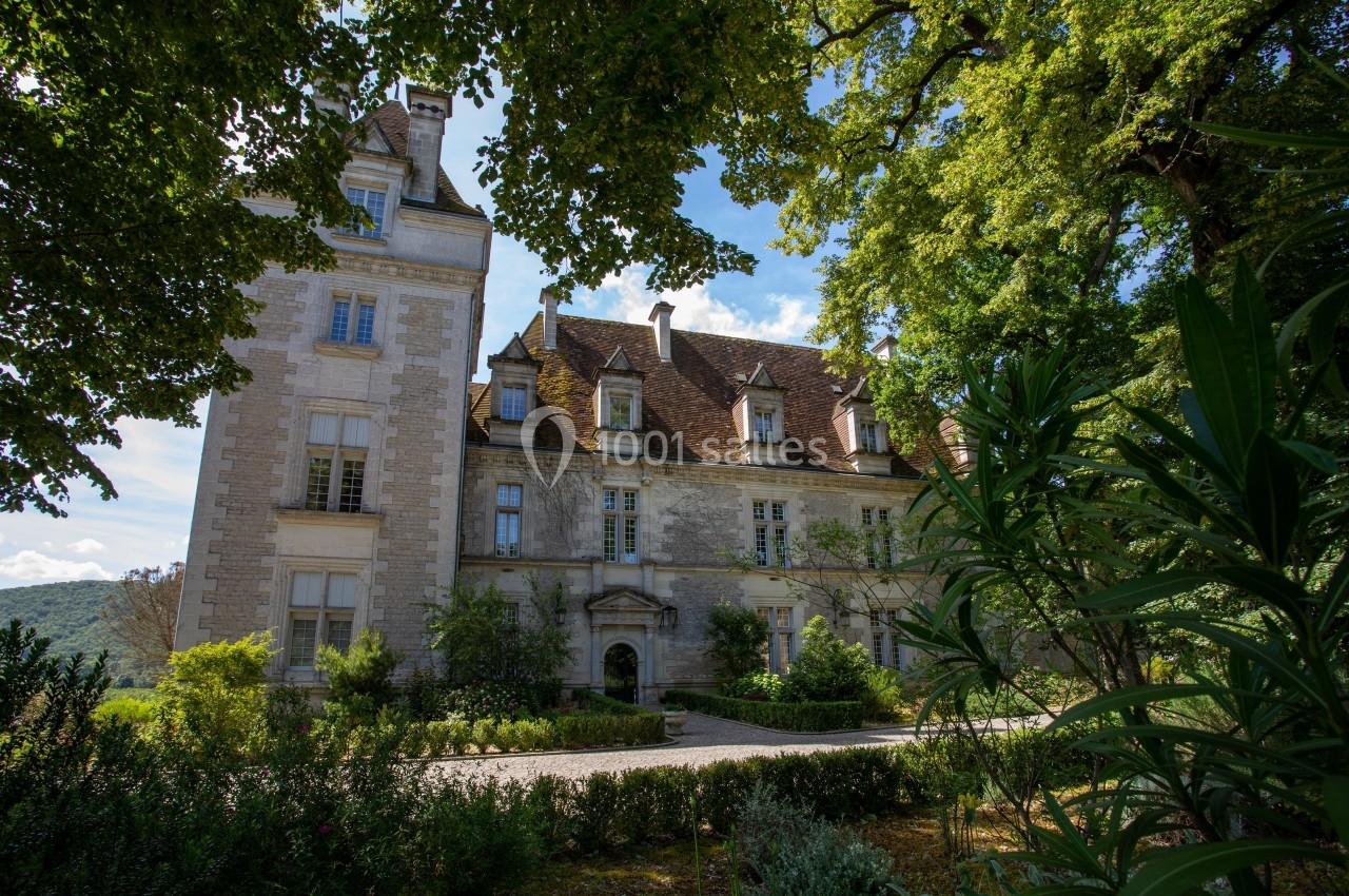 Façade d'un château en pierre entouré de végétation, avec un ciel bleu partiellement nuageux en arrière-plan.