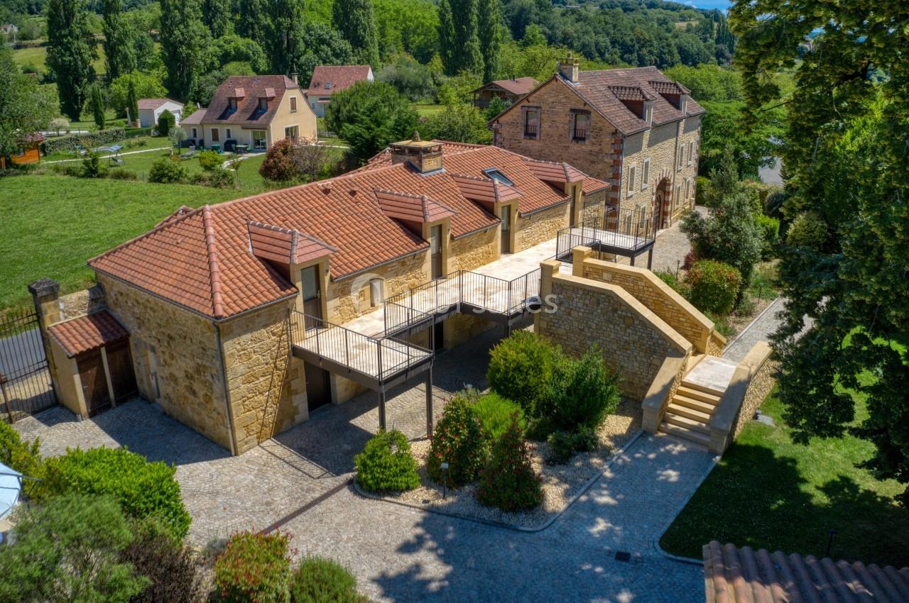 Vue aérienne d'un bâtiment en pierre avec toits en tuiles, entouré de verdure et d'autres habitations.