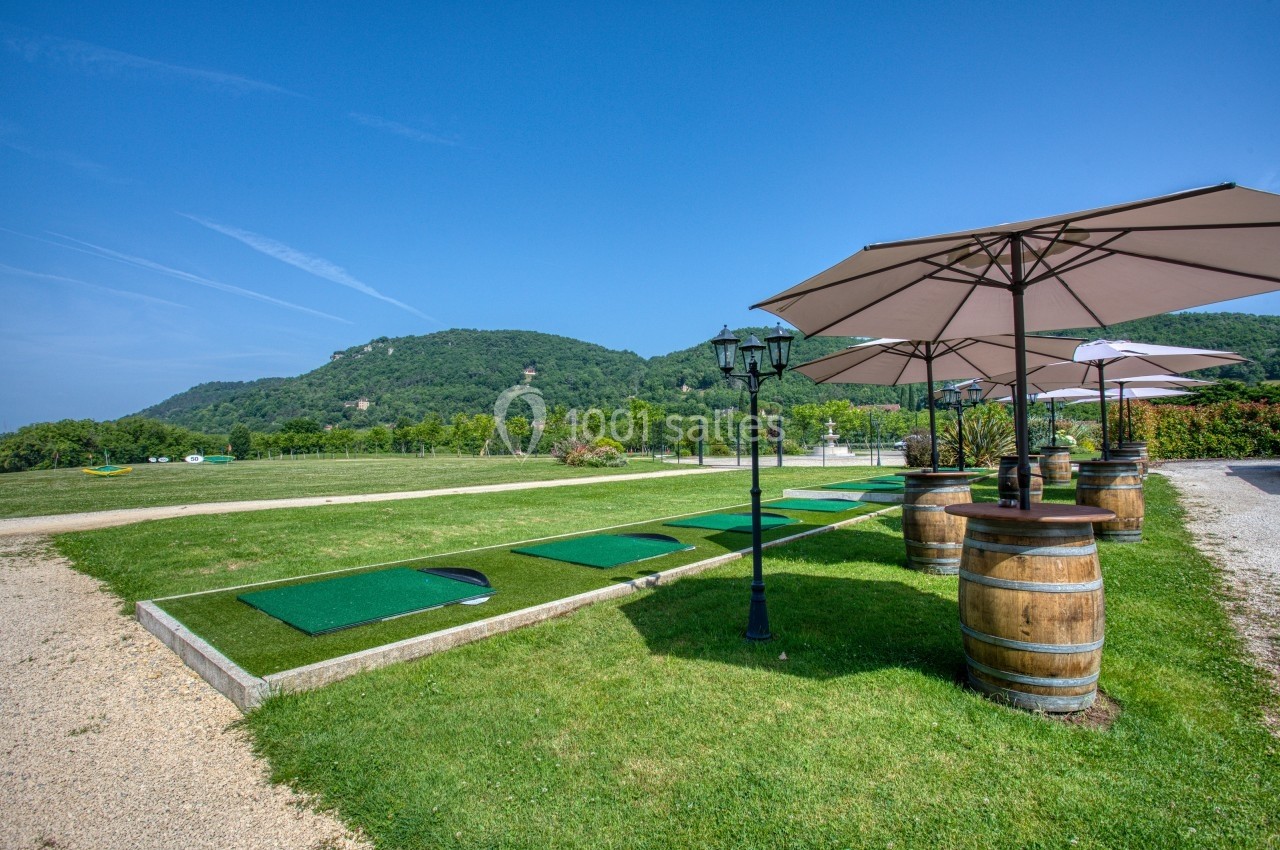 Terrains de practice de golf sur gazon avec parasols et tonneaux, entourés de collines verdoyantes sous un ciel bleu.