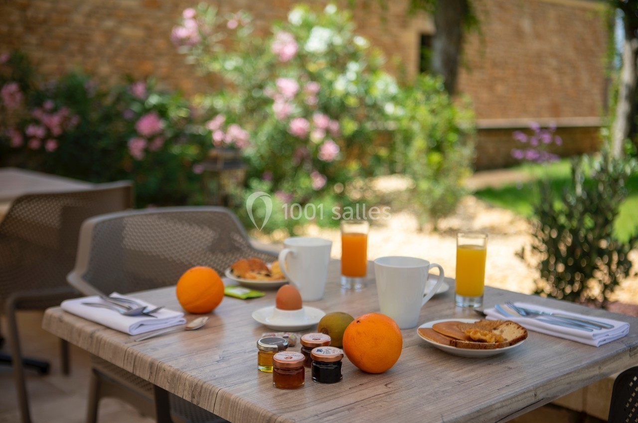 Petit-déjeuner en terrasse avec fruits, confitures, pain, œuf à la coque et jus d'orange, dans un jardin fleuri.
