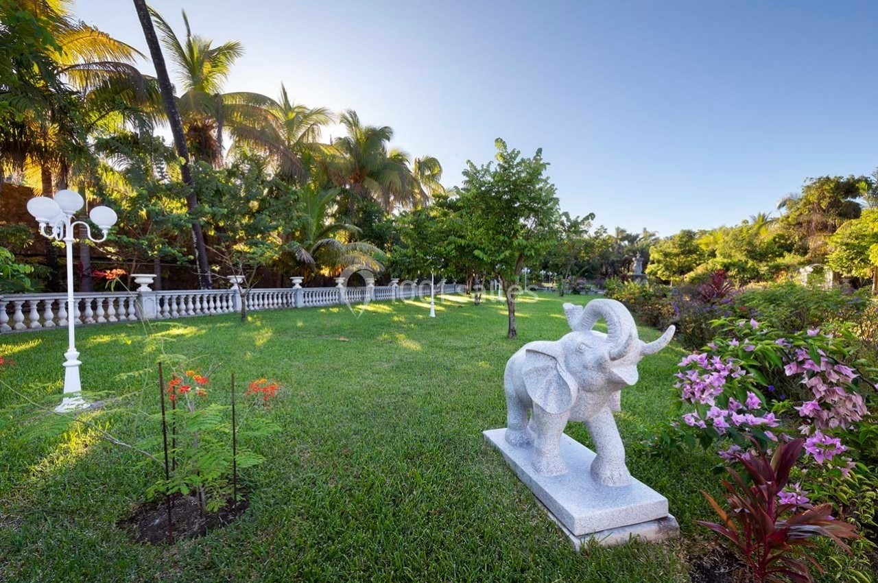 Statue d'éléphant en pierre dans un jardin verdoyant avec des fleurs, des palmiers et un lampadaire blanc.
