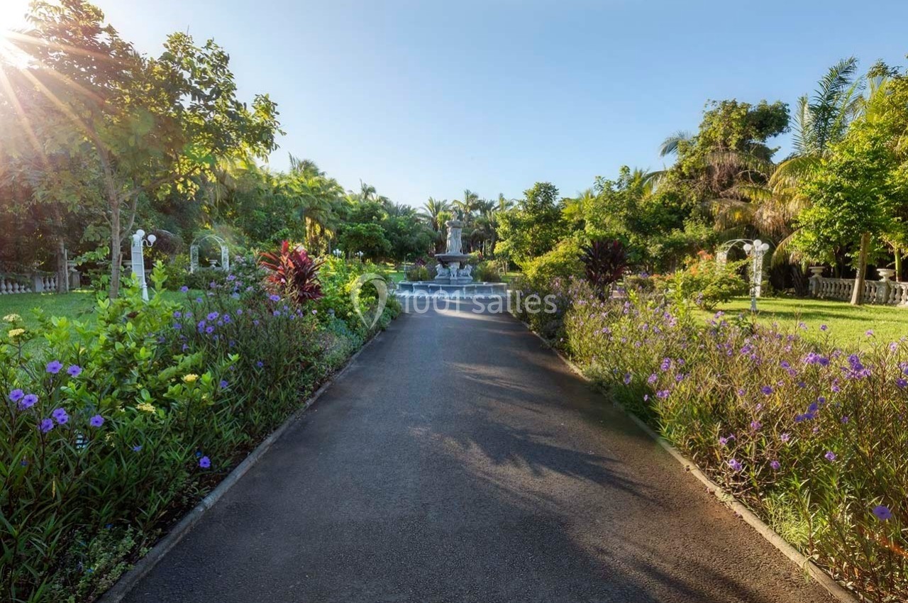 Allée bordée de fleurs et d'arbustes menant à une fontaine entourée de végétation luxuriante sous un ciel dégagé.
