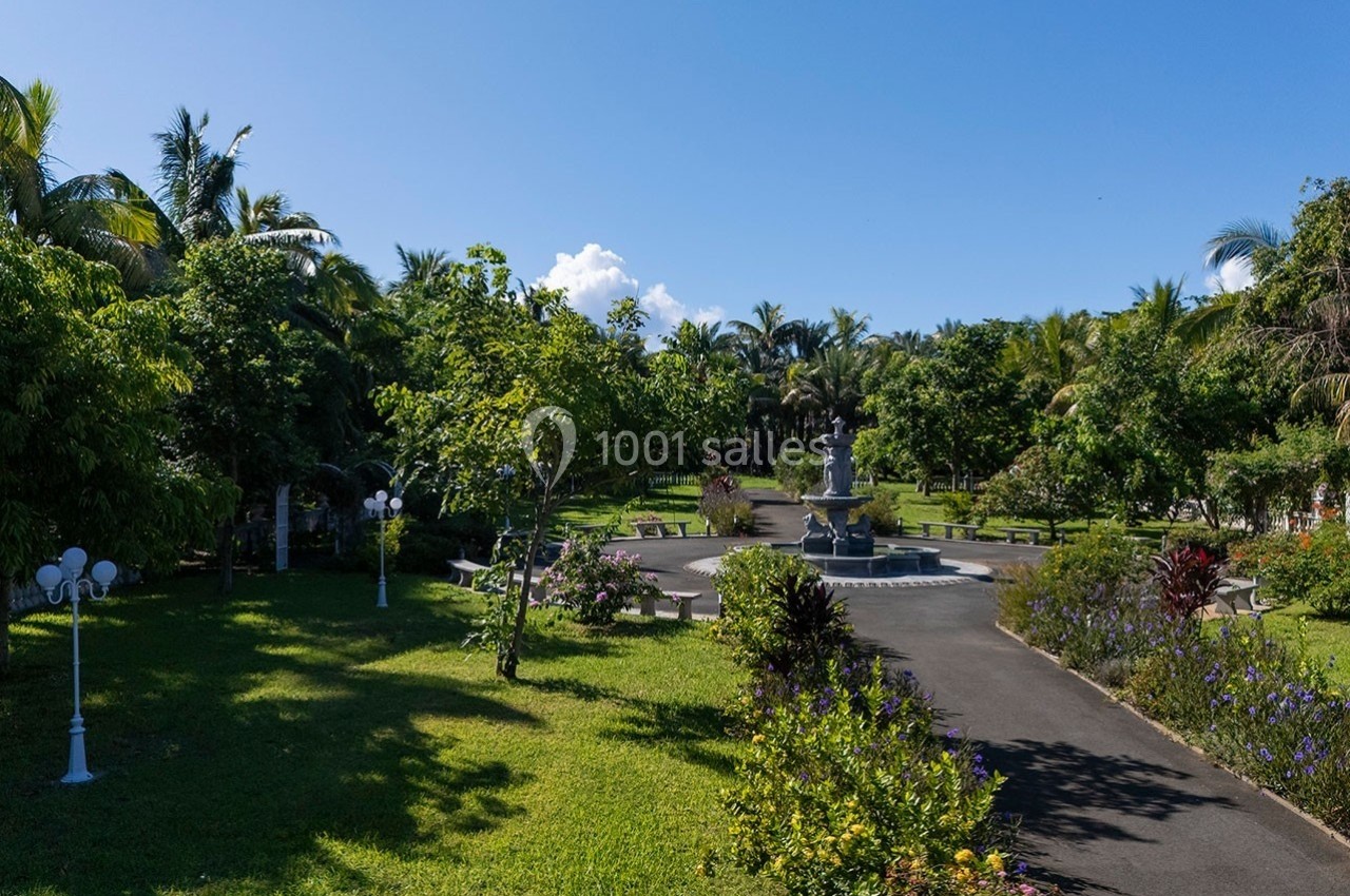 Allée bordée de végétation luxuriante avec une fontaine centrale entourée de bancs dans un parc ensoleillé.
