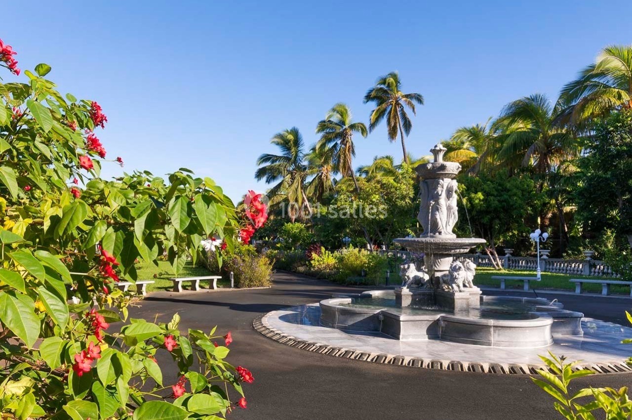 Fontaine en pierre entourée de végétation tropicale avec palmiers et fleurs rouges sous un ciel bleu.